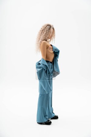 Photo for Full length of provocative and shirtless woman in blue oversize pantsuit covering naked breast with hand while standing on grey background, wavy ash blonde hair, passion and style, sexy look - Royalty Free Image