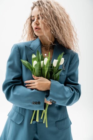 femininity and fashion, appealing and romantic woman with wavy ash blonde hair standing in blue oversize blazer and holding bouquet of white fresh tulips on grey background