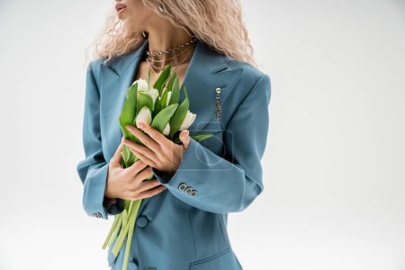 Teilansicht einer modischen Frau in blauem Oversize-Blazer mit Strauß weißer Tulpen auf grauem Hintergrund, welligem aschblondem Haar, silbernen Accessoires, modernem Stil