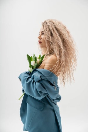 Photo for Side view of sexy woman with bare shoulders and wavy ash blonde hair wearing blue oversize blazer, holding bouquet of white tulips while standing and looking away on grey background, glamour style - Royalty Free Image