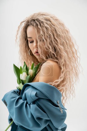 sinnliche und charmante Frau mit nackten Schultern und welligen aschblonden Haaren, die einen Strauß weißer Tulpen hält und in einem blauen Oversize Blazer auf grauem Hintergrund posiert, moderne Mode und Stil