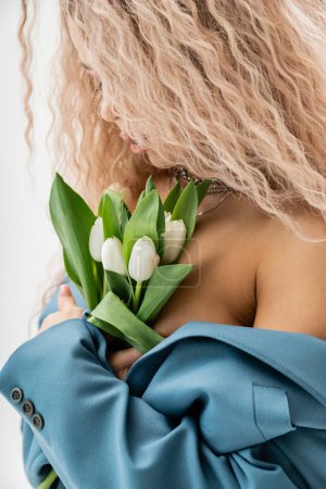 sexuality and fashion, charming and romantic woman with wavy ash blonde hair wearing blue oversize blazer on shirtless body and embracing bouquet of white tulips on grey background