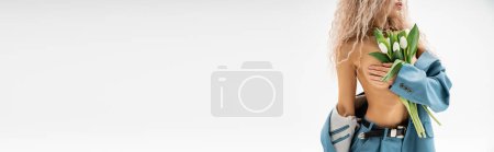 Foto de Vista recortada de mujer apasionada y sexy con cuerpo sin camisa quitándose la chaqueta azul, oscureciendo el pecho desnudo con la mano y sosteniendo tulipanes blancos sobre fondo gris, pancarta con espacio para copiar - Imagen libre de derechos