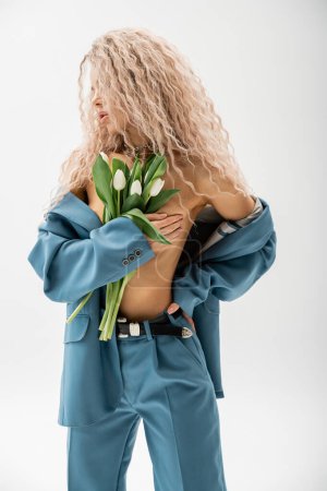 femme érotique avec des cheveux blonds cendrés ondulés portant blazer oversize bleu sur corps torse nu, couvrant poitrine nue avec la main et tenant bouquet de tulipes blanches sur fond gris, look sexy