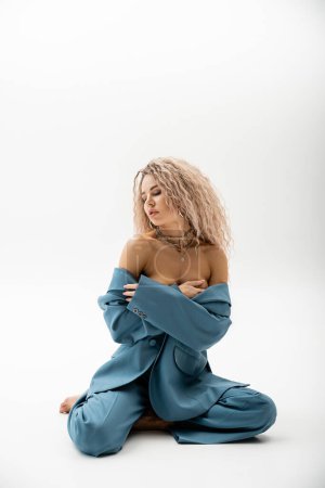 Photo for Full length of passionate woman with bare shoulders, wavy ash blonde hair and closed eyes sitting in blue oversize suit on grey background, sexy look, fashion and sensuality - Royalty Free Image