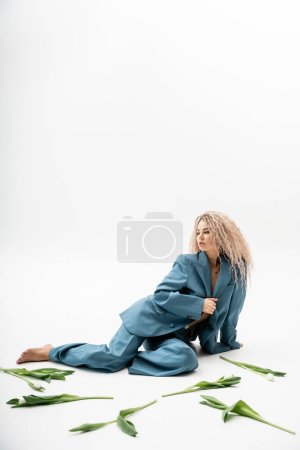 full length of sensual and charming barefoot woman with dyed ash blonde hair wearing blue oversize suit while sitting near tulips on grey background with copy space, modern individuality