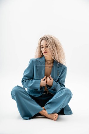 full length of expressive and fashionable barefoot woman in blue oversize suit sitting with crossed legs on grey background, wavy ash blonde hair, trendy look, sexuality and style