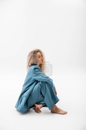 full length of dreamy and fashionable barefoot woman sitting on grey background and looking away, wavy ash blonde hair, blue oversize suit, sensuality and style, modern fashion photography