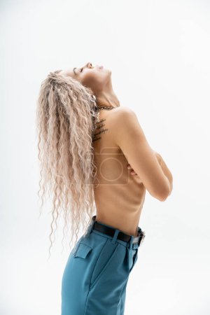 Photo for Side view of sensual woman with shirtless tattooed body and wavy ash blonde hair wearing blue oversize pants while standing with closed eyes on grey background, sexy look, self-expression - Royalty Free Image