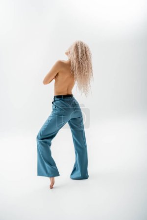 full length of sexy, shirtless and barefoot woman with dyed ash blonde hair posing in blue oversize pants on grey background, slender body, individuality and self-expression
