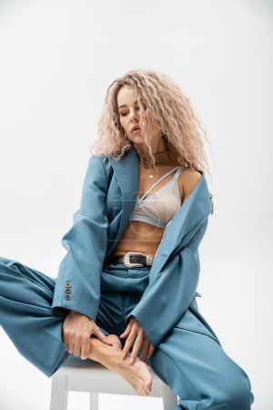 fashionable, barefoot and dreamy female model with wavy ash blonde hair, in bra and blue oversize suit posing on chair on grey background, sensual individuality, style and self-expression