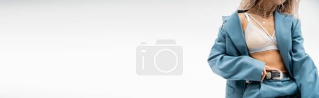 vista parcial de mujer seductora en collares de plata, sujetador y traje azul de gran tamaño tocando el cinturón de cuero mientras está de pie sobre fondo gris, aspecto sexy, moda moderna, pancarta con espacio para copiar