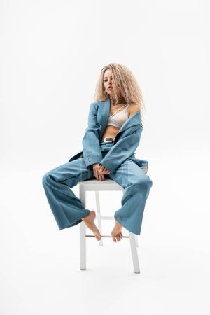 full length of sexy, appealing and trendy barefoot woman sitting on chair in bra and blue oversize suit on grey background, wavy ash blonde hair, sensual individuality, modern fashion