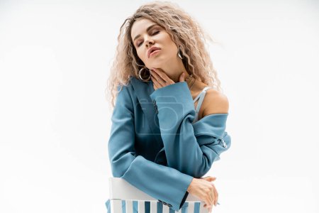 seductive and beautiful woman with wavy ash blonde hair and closed eyes touching neck and sitting on chair in blue oversize blazer and silver accessories on grey background