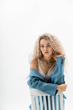 portrait of seductive woman in bra and blue oversize blazer sitting on chair and looking away on grey background, wavy ash blonde hair, expressive gaze, silver accessories, sexy fashion trend