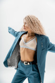 expressive and stylish woman with trendy silver accessories and wavy ash blonde hair wearing bra, blue oversize suit and posing with outstretched hands on grey background, sexuality and fashion mug #663916496