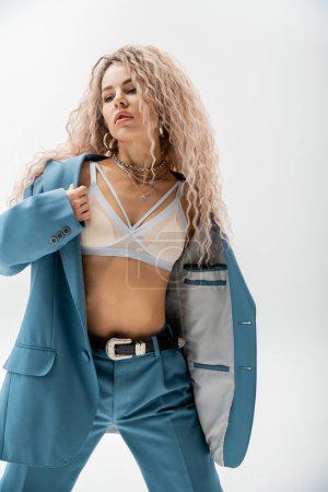 passionate woman in bra and blue oversize suit standing on grey background, wavy ash blonde hair, trendy silver accessories, slender body, sexy look, modern fashion photography