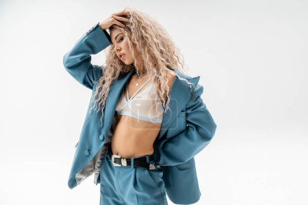 femininity and style, fashionable woman adjusting wavy ash blonde hair and holding hand behind back while posing in bra, silver necklaces and blue oversize suit on grey background