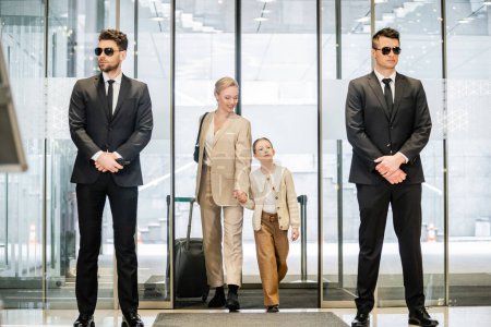 private security concept, two bodyguards in formal wear and sunglasses standing near hotel entrance, cheerful woman and child holding hands and walking with luggage, entering lobby, luxury lifestyle 