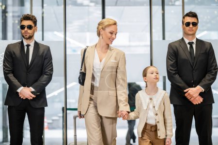 Photo for Private security service, two bodyguards in formal wear and sunglasses standing near hotel entrance, happy woman and child holding hands and walking with luggage, entering lobby, luxury lifestyle - Royalty Free Image