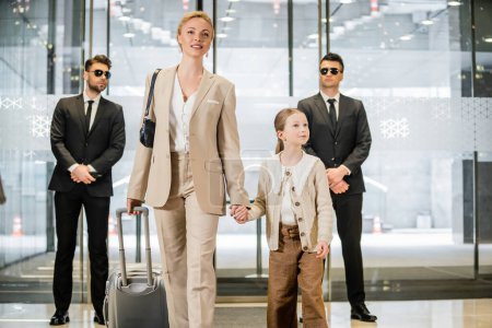 Photo for Personal security and protection, two bodyguards in suits and sunglasses standing near hotel entrance, happy mother and child holding hands and walking with luggage, entering lobby, luxury lifestyle - Royalty Free Image