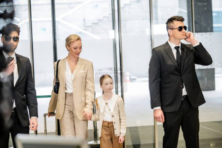 personal security and protection, two bodyguards in suits and sunglasses communicating while standing near hotel entrance, happy mother and child entering lobby, luxury lifestyle 