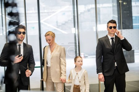 personal security concept, two bodyguards in suits and sunglasses protecting happy mother and child entering lobby, luxury lifestyle, woman and girl holding hands and walking inside 