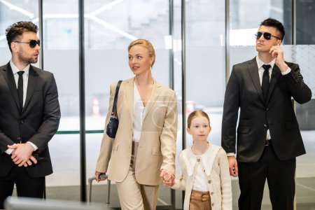 personal security concept, two bodyguards in suits and sunglasses protecting happy blonde woman and child, luxury lifestyle, woman and girl holding hands and walking inside of hotel 