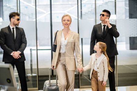 luxury lifestyle, personal security concept, two bodyguards in suits and sunglasses protecting attractive blonde woman with child, woman and girl holding hands and walking inside of hotel 