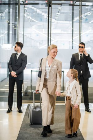Photo for Luxury lifestyle, rich family, personal security concept, two bodyguards in suits and sunglasses protecting beautiful blonde woman with child, woman and girl holding hands and walking inside of hotel - Royalty Free Image