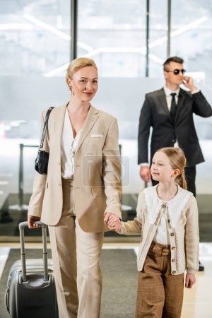 luxury lifestyle, rich, personal security, happy woman with child, blonde woman and girl holding hands and walking inside of hotel, bodyguard in suit and sunglasses standing on blurred background 