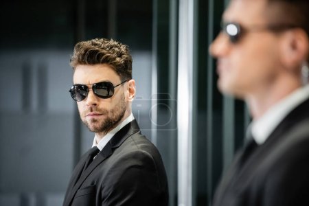 bodyguard service, private security, handsome man in suit and sunglasses standing in hotel lobby near work partner, vigilance, protection and career, professional headshots, looking at camera  