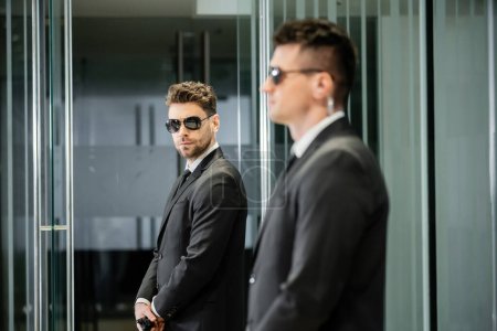 Bodyguard-Service, Hotelsicherheit, gutaussehender Mann in Anzug und Sonnenbrille, der in der Lobby in der Nähe seines Arbeitspartners steht, Wachsamkeit, Schutz und Arbeit, professionelle Kopfschüsse, Blick in die Kamera  