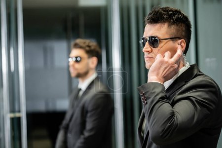 Photo for Bodyguard service, private security, professional guards in suits and sunglasses standing in hotel lobby, handsome man with earpiece communicating with work partner, luxury hotel, vigilance - Royalty Free Image
