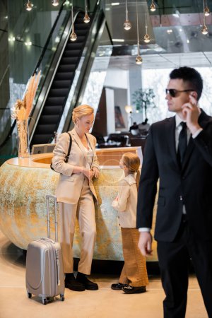 personal safety concept, blonde woman standing at reception desk with preteen daughter, rich lifestyle, family travel, bodyguard in suit and sunglasses standing on blurred foreground, safety