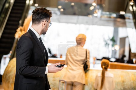 bodyguard concept, handsome man in suit and tie holding radio transceiver, protecting clients on blurred background, looking at mother and daughter at reception desk, connection and safety