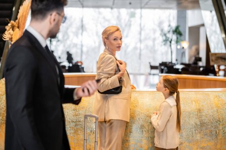 personal security, lifestyle, blonde mother with child standing at reception desk, woman and preteen girl looking at camera during check in, bodyguard in suit standing on blurred foreground 