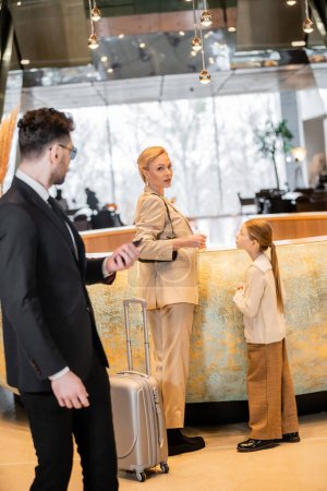 personal security, lifestyle, blonde mother with preteen girl standing at reception desk, successful woman looking at camera during check in, bodyguard in suit standing on blurred foreground 