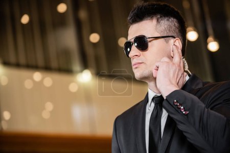 handsome bodyguard in sunglasses, handsome man in suit and tie touching earpiece in lobby of hotel, security, career, communication, vigilance, private safety, hotel staff 