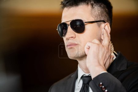 Photo for Handsome bodyguard in sunglasses, handsome man in suit and tie touching earpiece in lobby of hotel, security, career, communication, vigilance, private safety, hotel industry, male personnel - Royalty Free Image