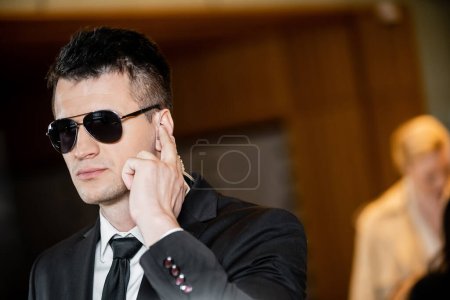 gutaussehender Bodyguard mit dunkler Sonnenbrille, gutaussehender Mann in Anzug und Krawatte, der Ohrhörer in der Lobby des Hotels berührt, Sicherheit und Karriere, Kommunikation, Wachsamkeit, private Sicherheit, Hotelsicherheit, männliches Personal 