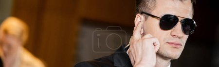 handsome bodyguard in sunglasses, man in suit and tie touching earpiece in lobby of hotel, security, career, communication, vigilance, private safety, hotel safety, male personnel, banner