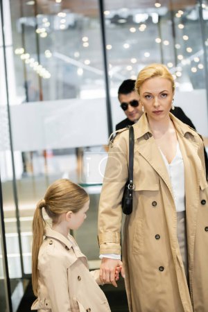 Photo for Private security service, bodyguard in formal wear and sunglasses standing near hotel entrance, blonde woman and child entering hotel lobby, autumn fashion, warm clothing, luxury lifestyle - Royalty Free Image