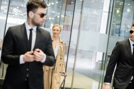 private security, blonde woman in trench coat entering into hotel, walking with luggage, two bodyguards in formal wear and sunglasses on blurred foreground, personal safety 