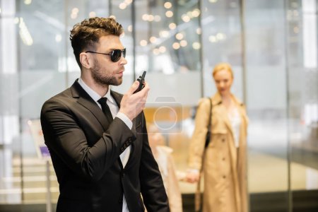 Bodyguard-Konzept, gutaussehender Mann in Anzug und Krawatte mit Funkgerät, schützt Kunden auf verschwommenem Hintergrund, spricht mit Walkie Talkie, Verbindung und Sicherheit