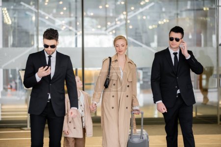 Persönliches Sicherheitskonzept, zwei Leibwächter in Anzügen und Sonnenbrillen schützen blonde Frau mit Kind beim Betreten des Hotels mit Gepäck, tragen herbstliche Kleidung, Luxus und reichen Lebensstil 