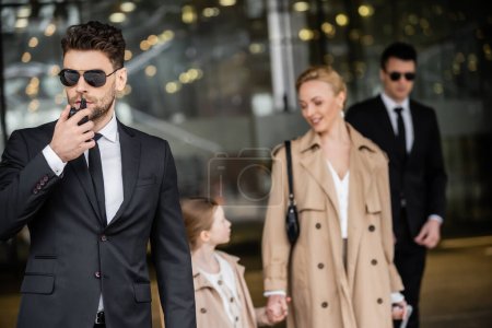 handsome bodyguard communicating through walkie talkie, standing next to successful woman and preteen kid, private security, blonde mother and daughter in trench coats, safety and protection 