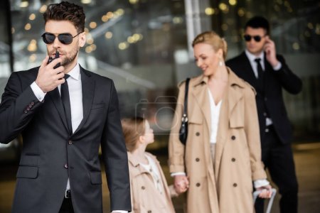 Photo for Handsome bodyguard communicating through walkie talkie, standing next to successful woman and preteen child, private security, blonde mother and daughter in trench coats, safety and protection - Royalty Free Image