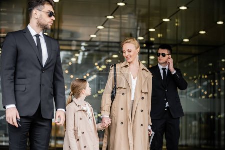 two bodyguards protecting successful woman and preteen kid, private security, happy blonde mother and daughter in trench coats near hotel, safety and protection, family travel, rich life 