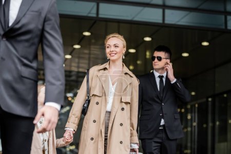 professional bodyguards protecting smiling blonde woman and preteen kid, successful mother and daughter in trench coats standing near hotel, safety and protection, private security, guards 